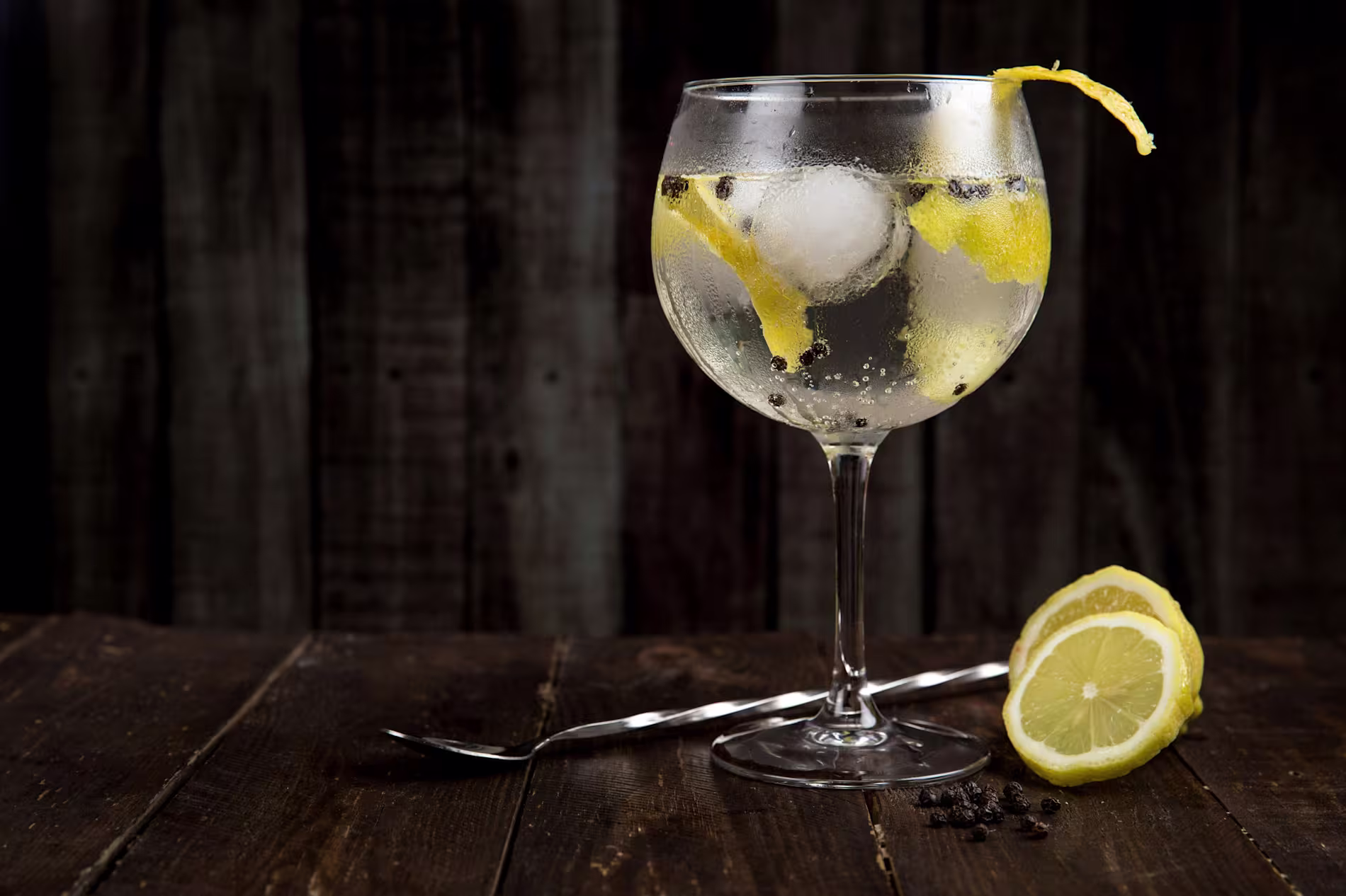 A gin and tonic with ice, lemon peel, and peppercorns in a large glass sits on a wooden table next to a twisted spoon and lemon slices.