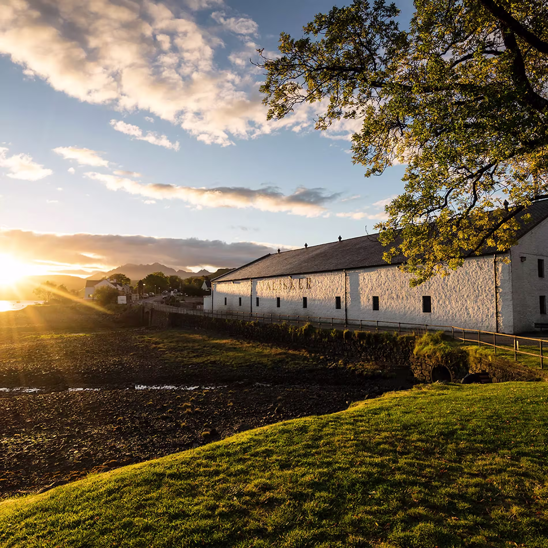 White stone building beside a rocky shoreline at sunset, with sunlight casting long shadows across grass and trees in the foreground.