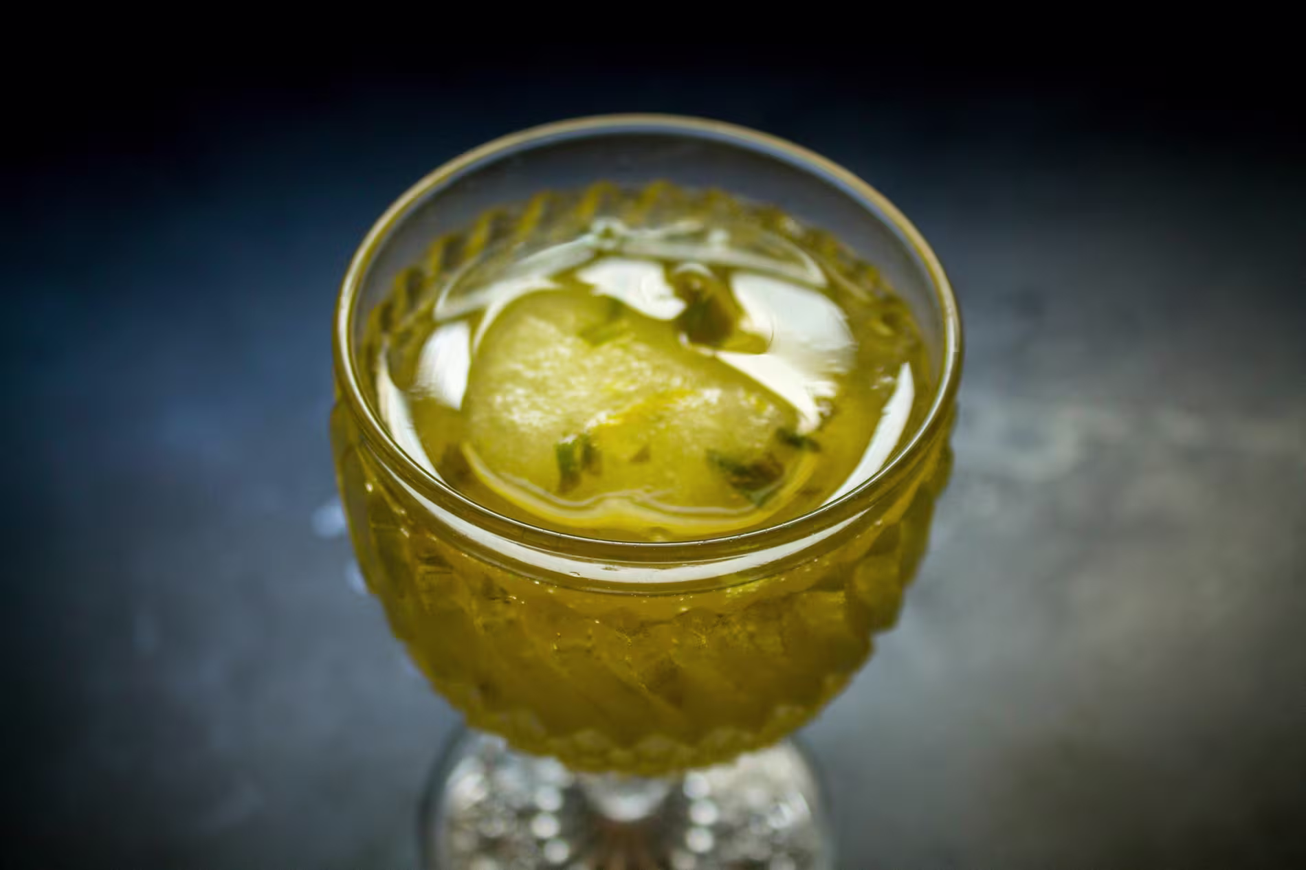 A close-up of a yellow drink with ice in a decorative glass on a dark surface.