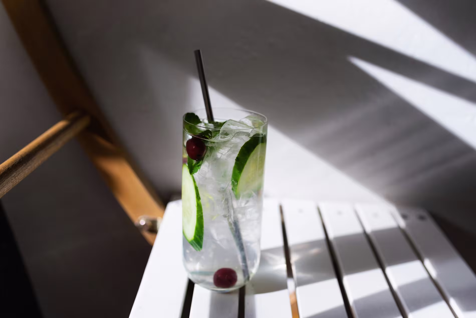 A glass of ice water with cucumber slices, mint leaves, and red berries on a white slatted table, with sunlight casting shadows.