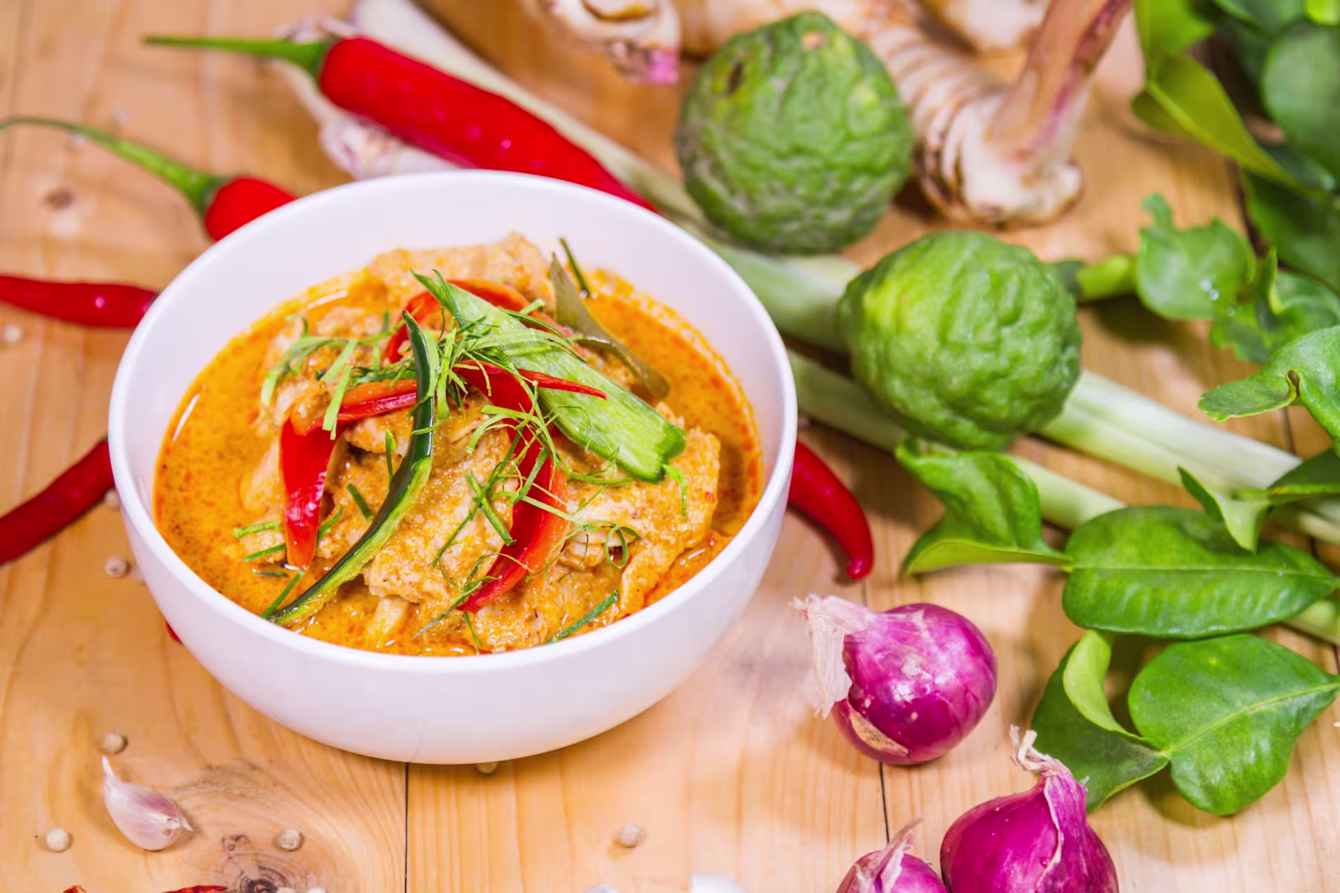 A bowl of Thai red curry with meat and vegetables, garnished with red chili, surrounded by fresh ingredients like lime, chili peppers, lemongrass, and shallots on a wooden surface.