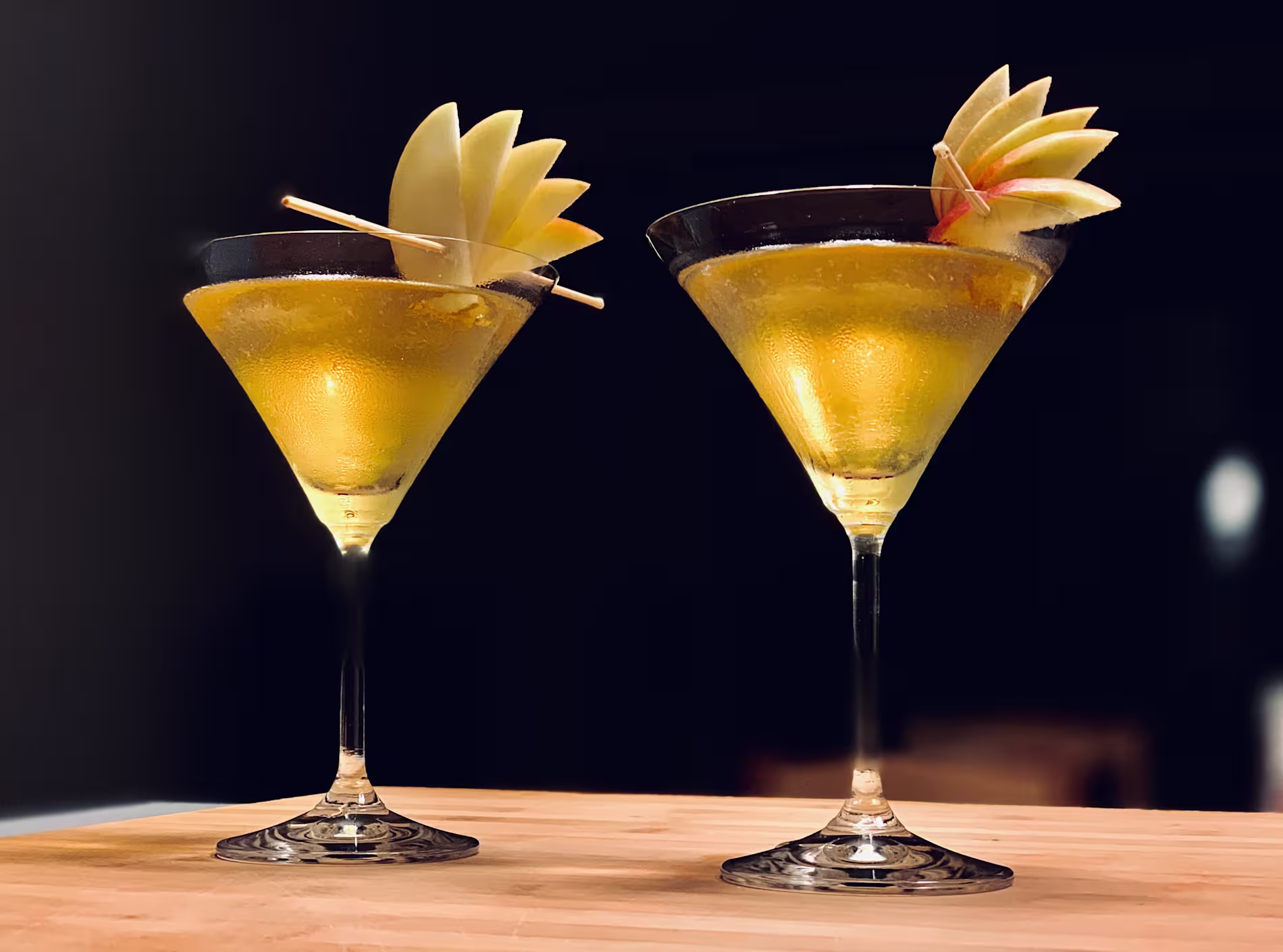 Two martini glasses filled with a golden cocktail, each garnished with a fan of thin apple slices and a red cocktail pick, placed on a wooden surface against a dark background.