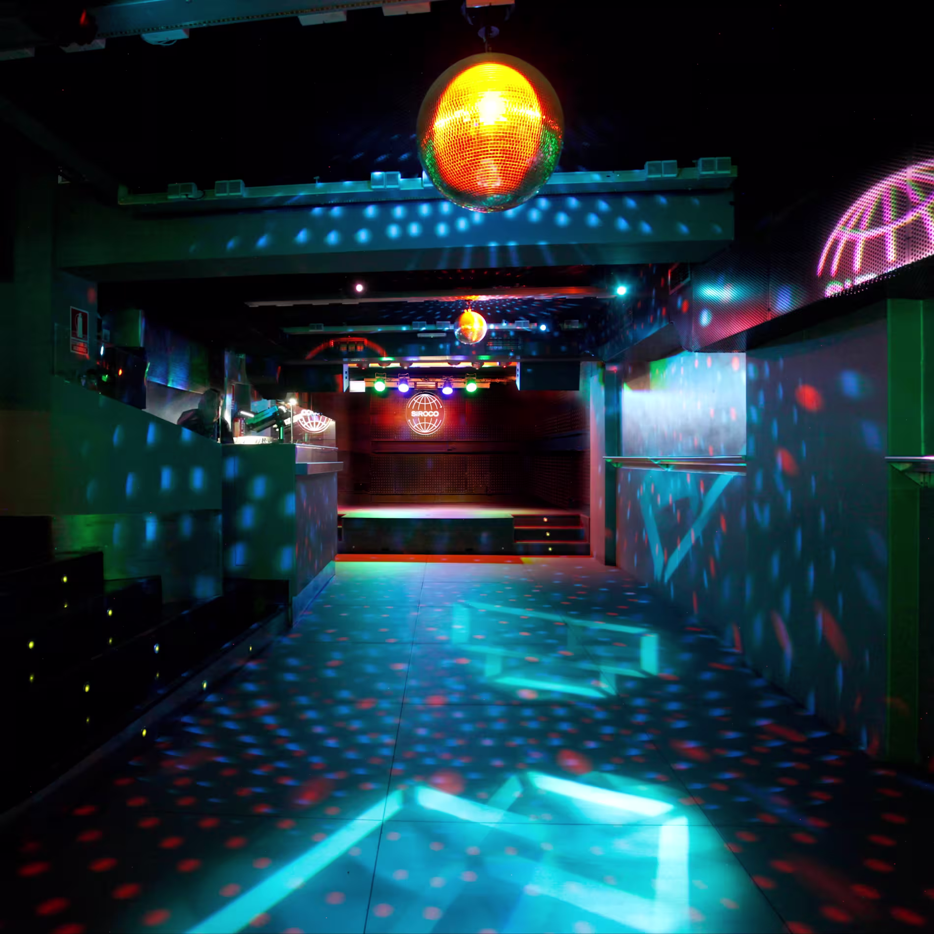 La foto muestra una pista de baile de la sala sirocco con efectos de iluminación coloridos. Hay luces azules y verdes proyectadas en el suelo, creando patrones, y una bola de discoteca colgando del techo que proyecta puntos de luz alrededor de la sala. 
