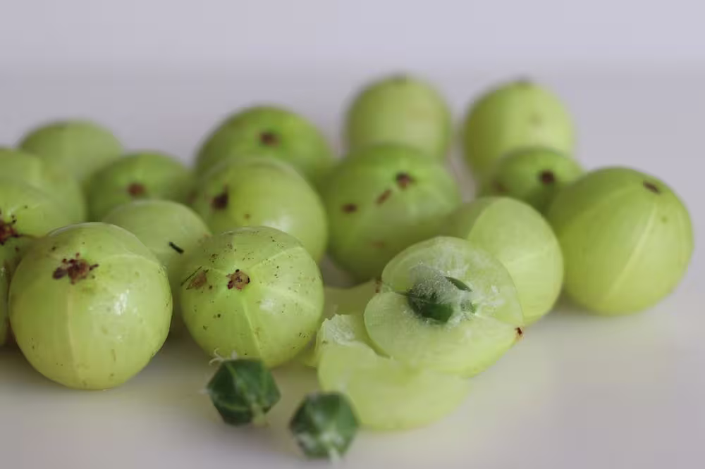 Several fresh Indian gooseberries (amla) are scattered on a white surface, with one fruit cut open to show its inner seed.