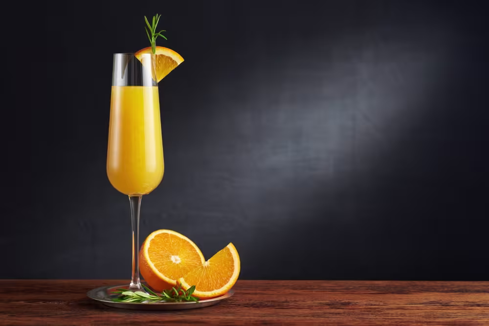 A glass of orange juice garnished with a rosemary sprig and orange slice, placed on a tray with orange wedges and greenery on a wooden surface against a dark background.