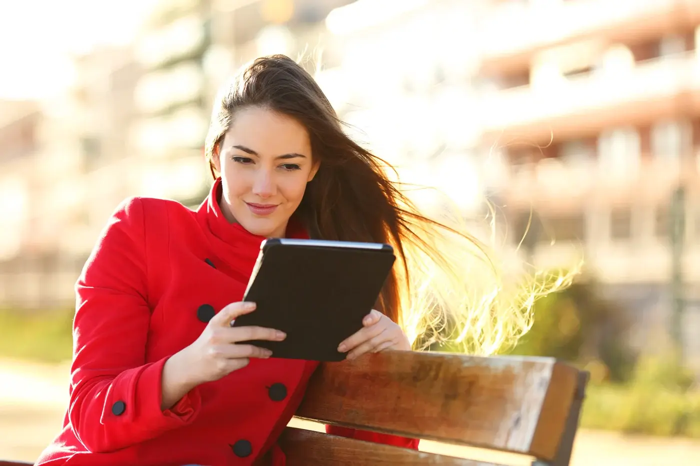 Woman in a red coat reading a tablet outdoors on a sunny day
