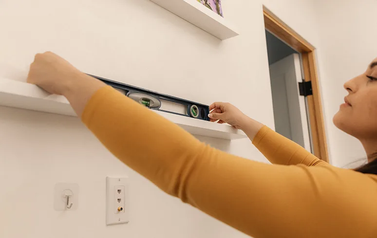 Woman in an orange shirt measuring a white shelf with a level measuring tool on top of a white wall.