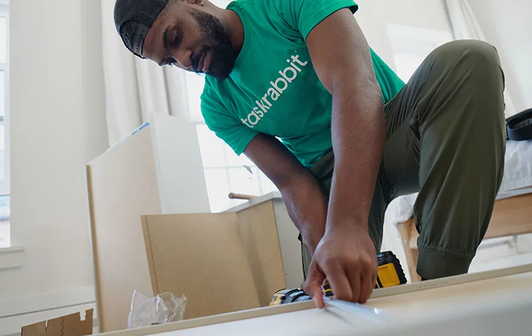 Man in a green Taskrabbit shirt crouching over a furniture piece with a yellow and black cordless drill driver.