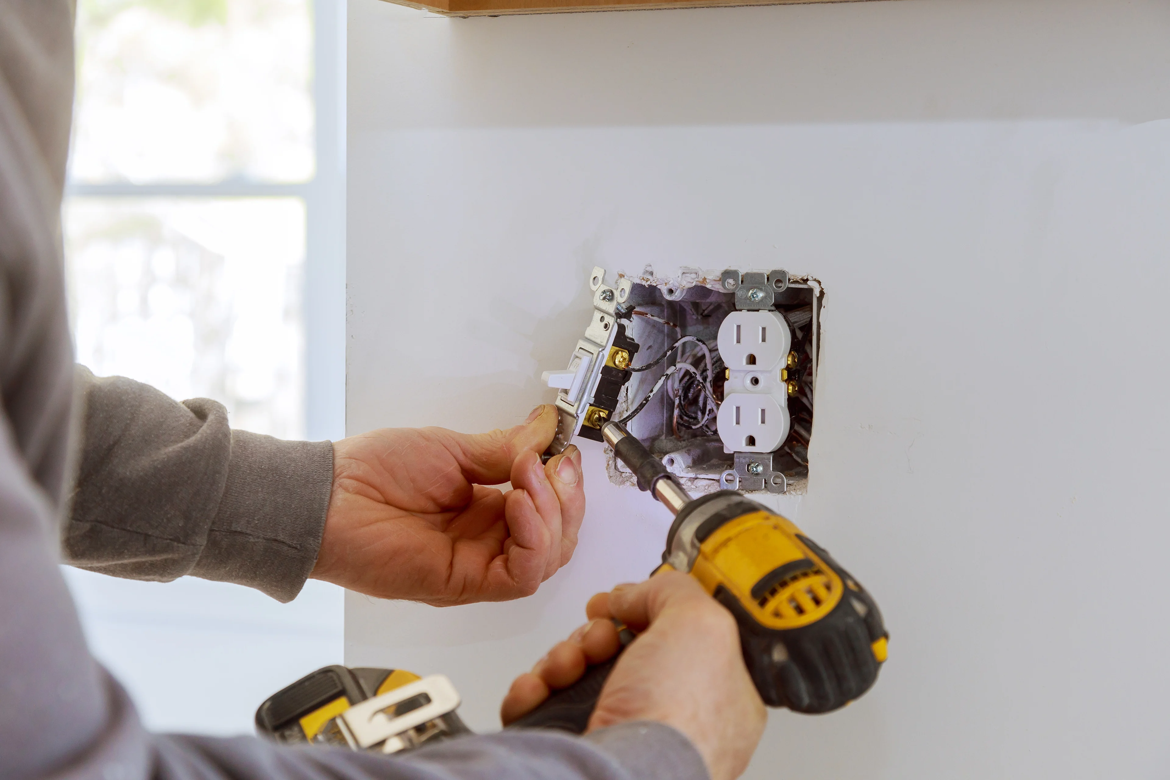 Una persona instalando un interruptor con un taladro junto a una toma de corriente. 