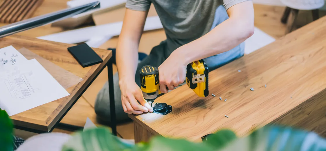 A picture of some onw using a drill to assemble a table. 