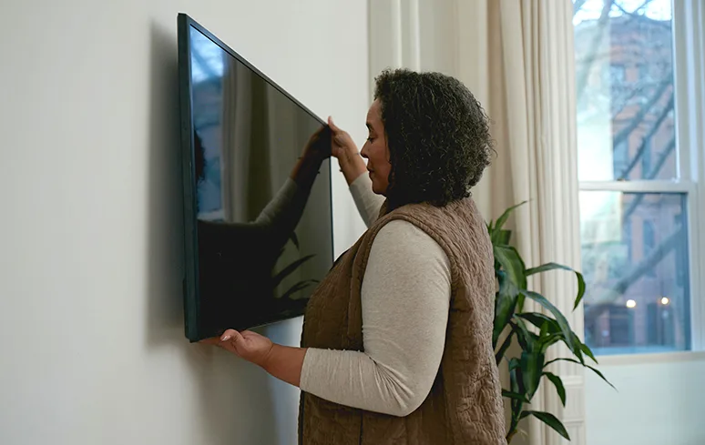 Woman in a brown shirt hanging a black flat screen TV on a white wall next to a floor plant and white windows.