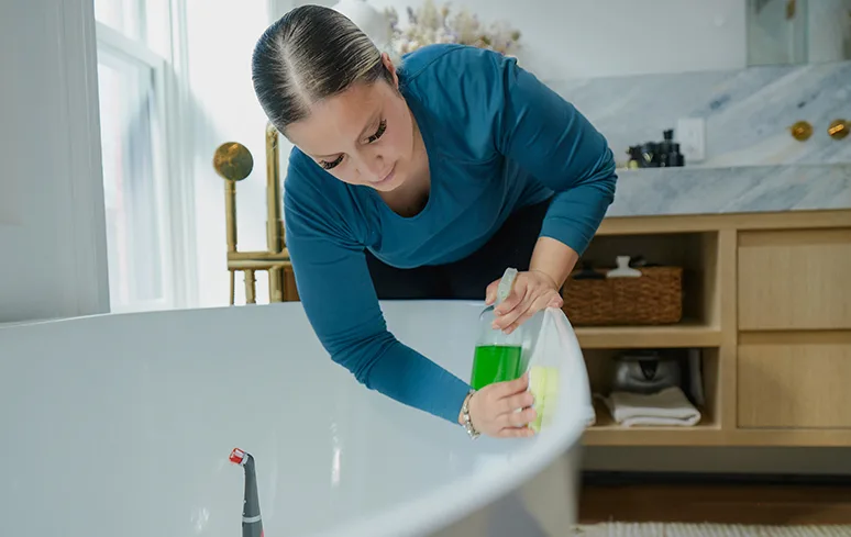 Woman in a blue shirt leaning over a white tub cleaning it with a sponge and a green cleaning spray bottle in a bathroom.