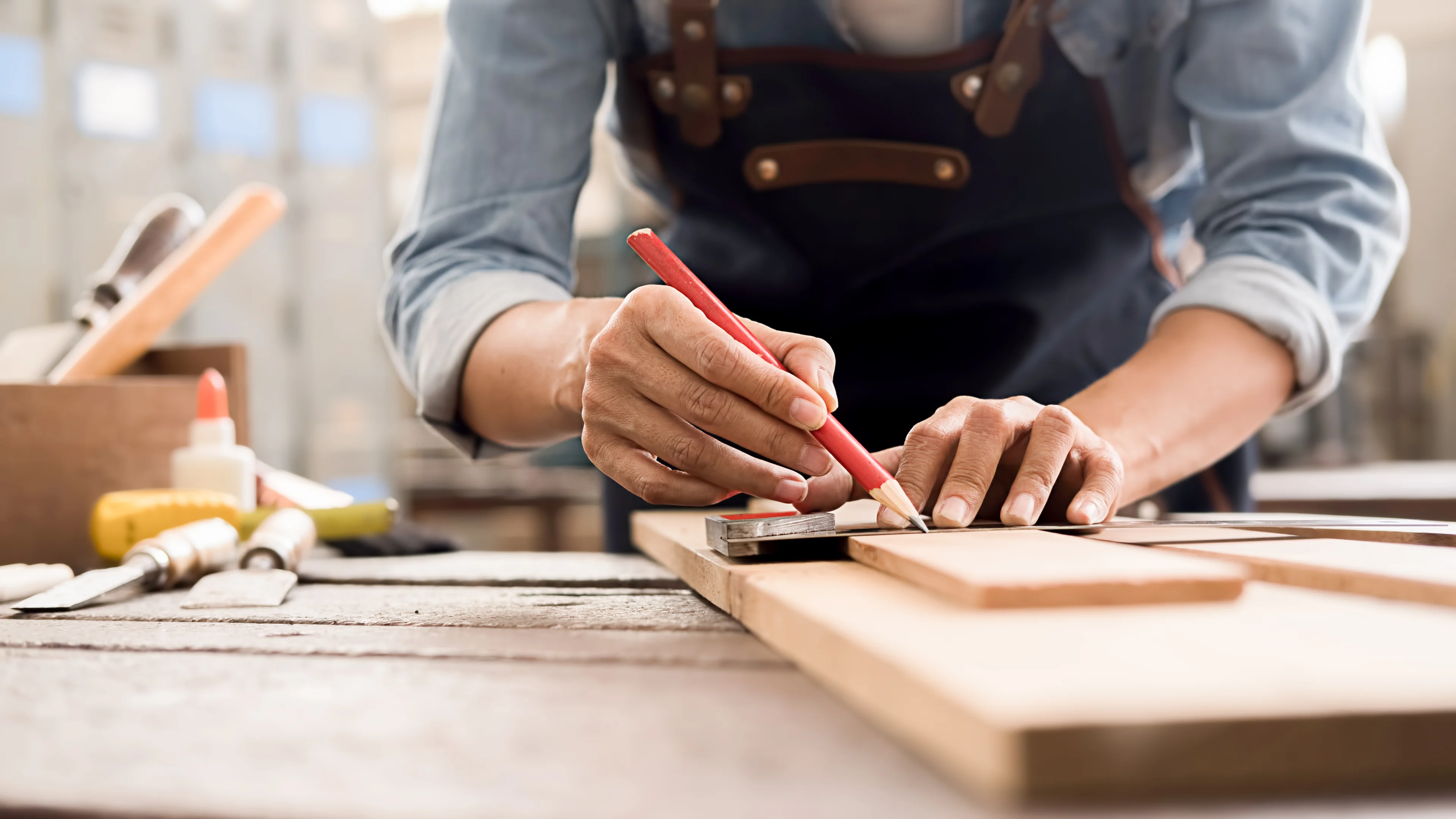 A carpenter wearing an apron marking measurements on a piece of wood inside of a wood working shop