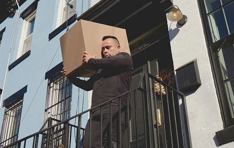 Man dressed in black holding a big cardboard box on one shoulder as he walks down stairs outside a house.