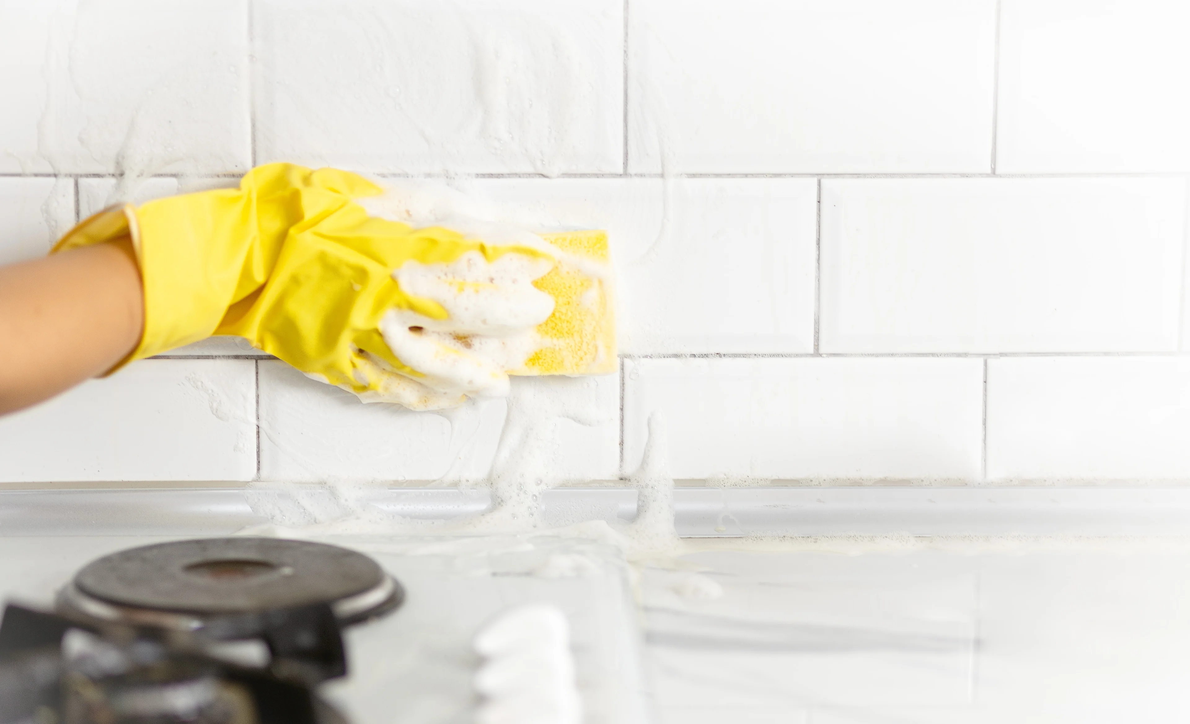 a person scrubbing backsplash tile behind a stove with a sponge and gloves. 