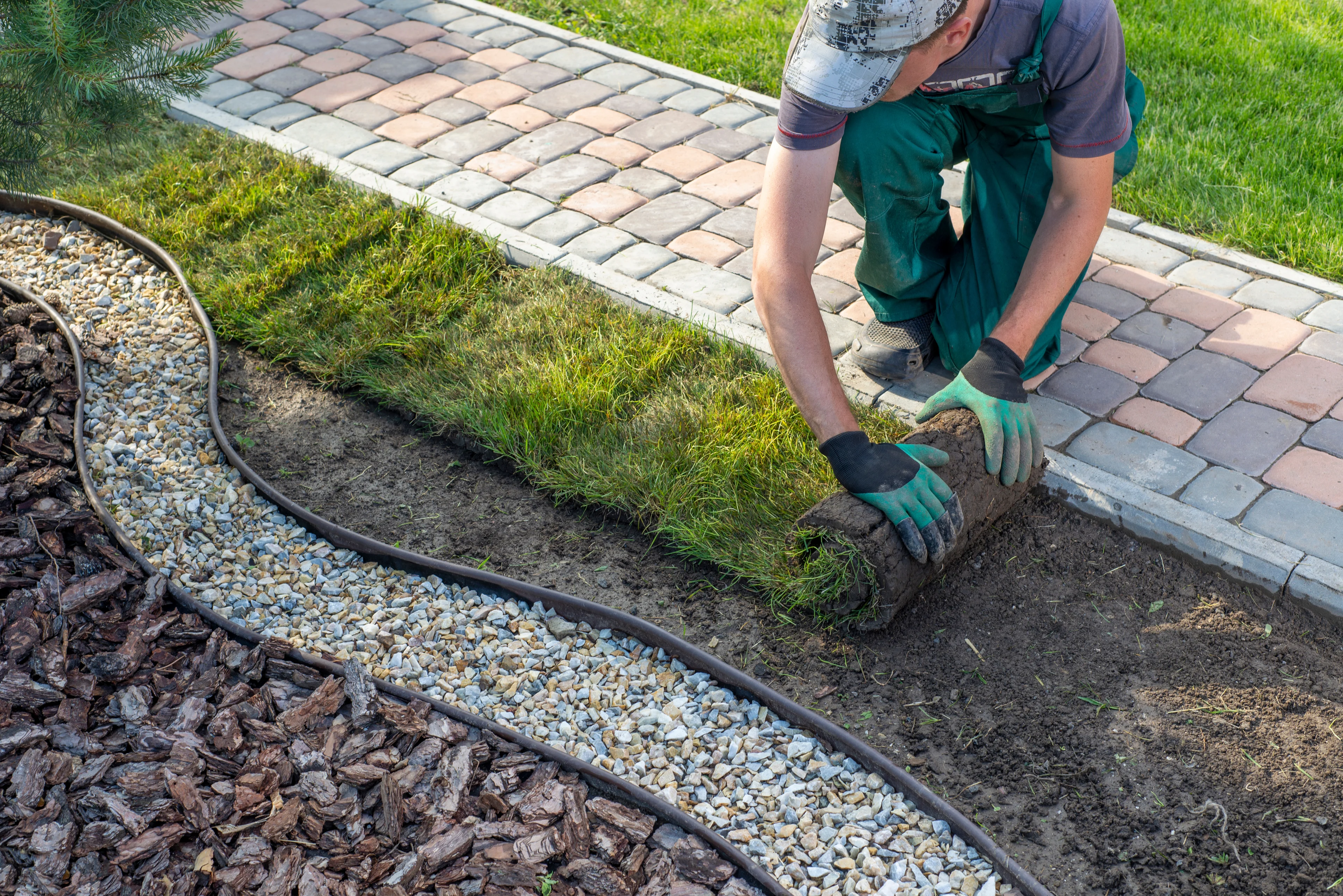 person laying down sod near a flowerbed and stone path in the yard of a home. 