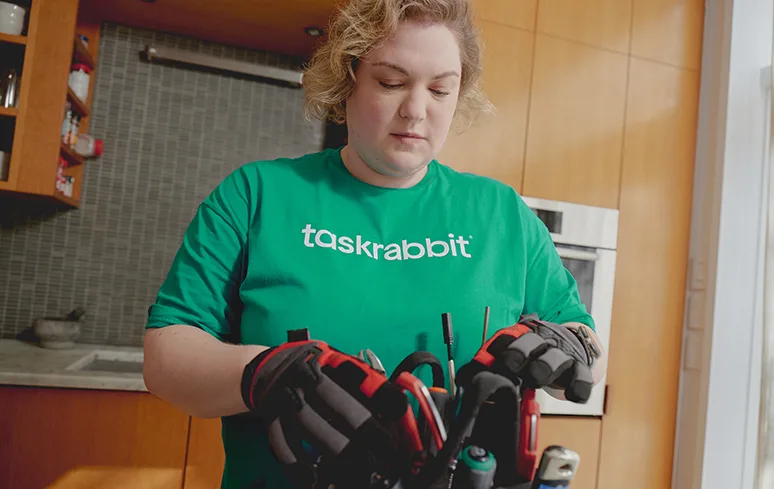 Woman in a green Taskrabbit shirt with gloves looking down at a toolkit.