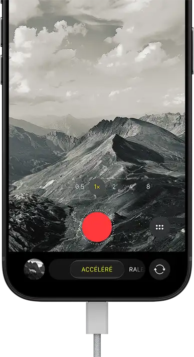 Un iPhone 18 Pro, face avant, affichant un paysage montagneux, avec le menu superposé du mode caméra en mode accéléré, câble d’alimentation branché au port de recharge