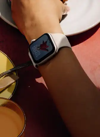 Le bras d’une personne sur la table à déjeuner, Apple Watch SE 3 sur son poignet, boîtier en aluminium comète, écran toujours allumé, portrait d’une personne sur le cadran, bracelet sport comète