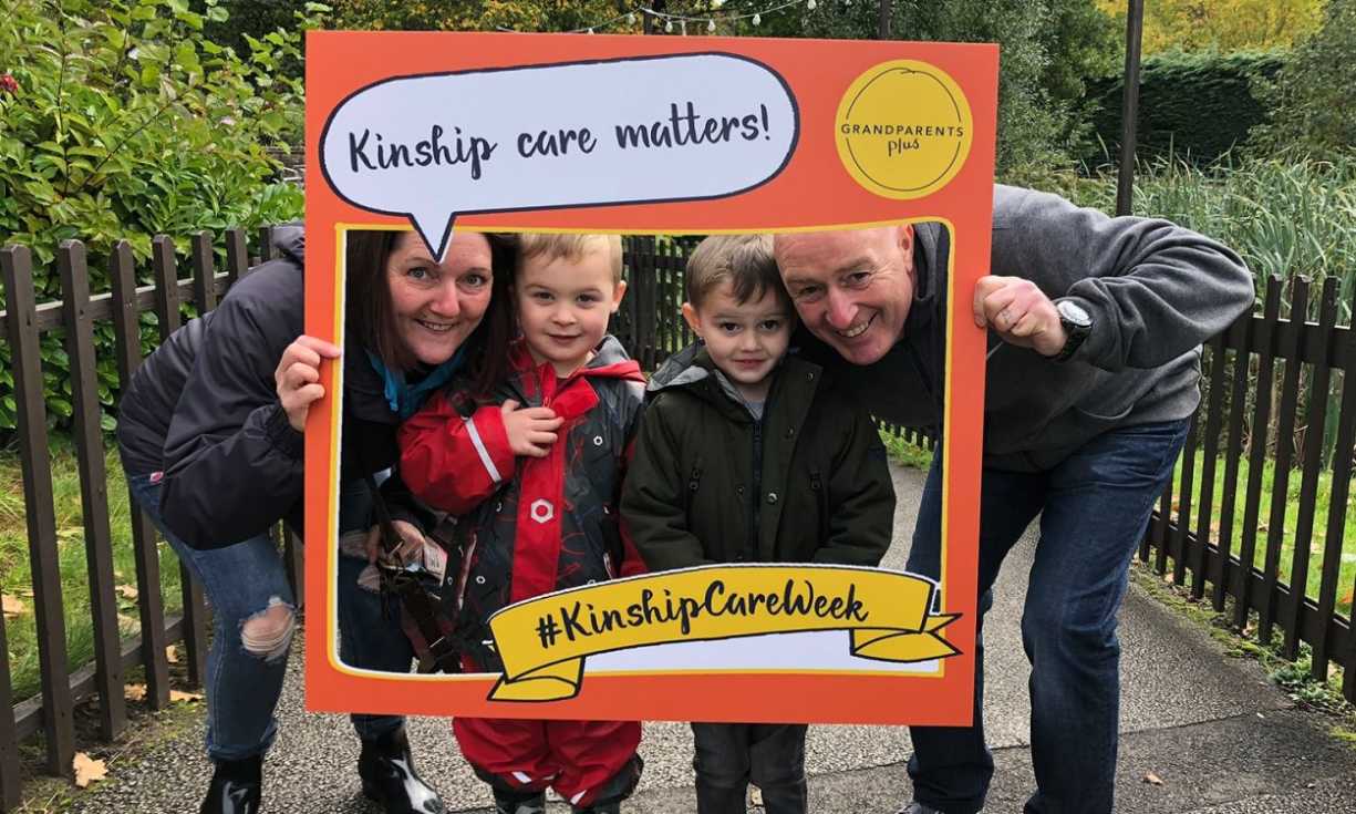 A man and woman, accompanied by two children, with a Grandparents Plus #KinshipCareWeek photo frame