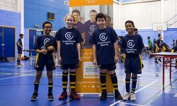 Greenhouse Sports. Four children in purple Greenhouse Sports t-shirts posing in a gym hall with ping-pong tables