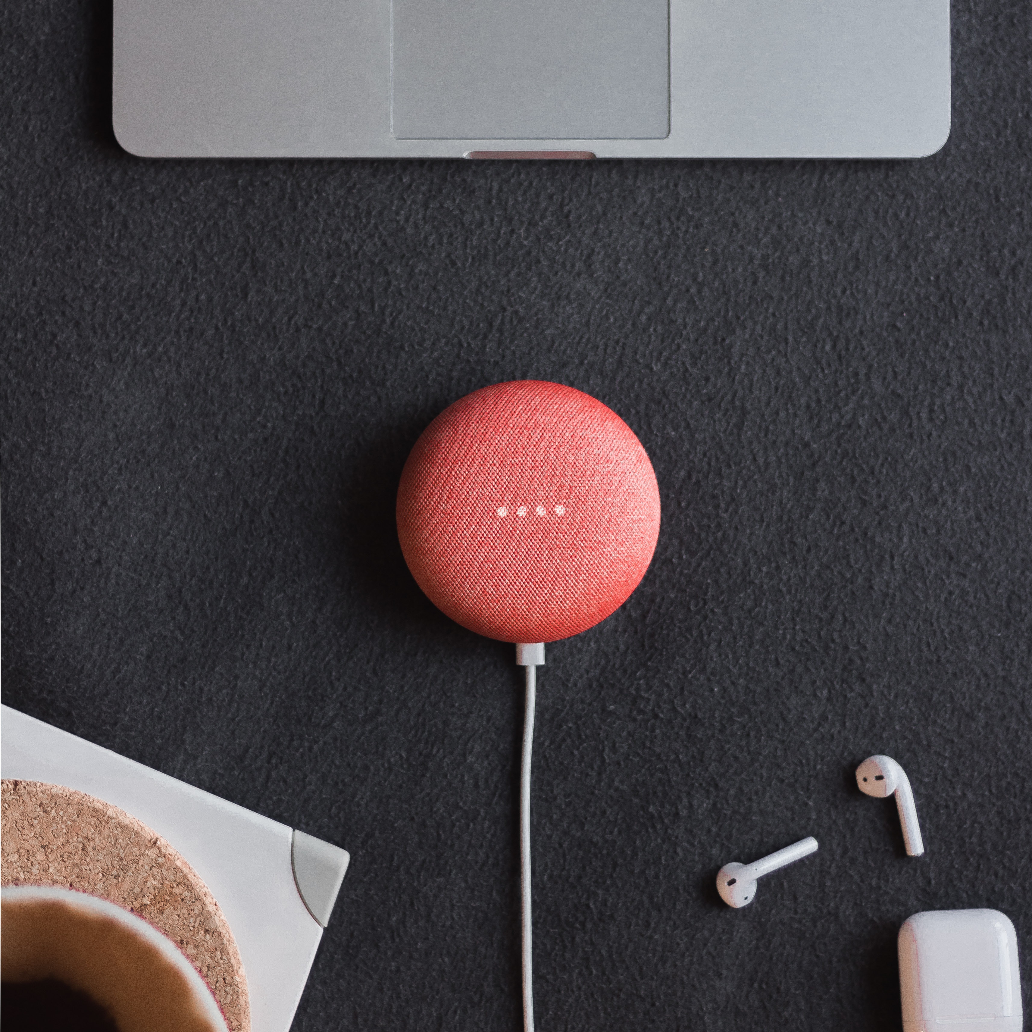 A red Google Home Mini surrounded by a pair of Apple AirPods and a MacBook.