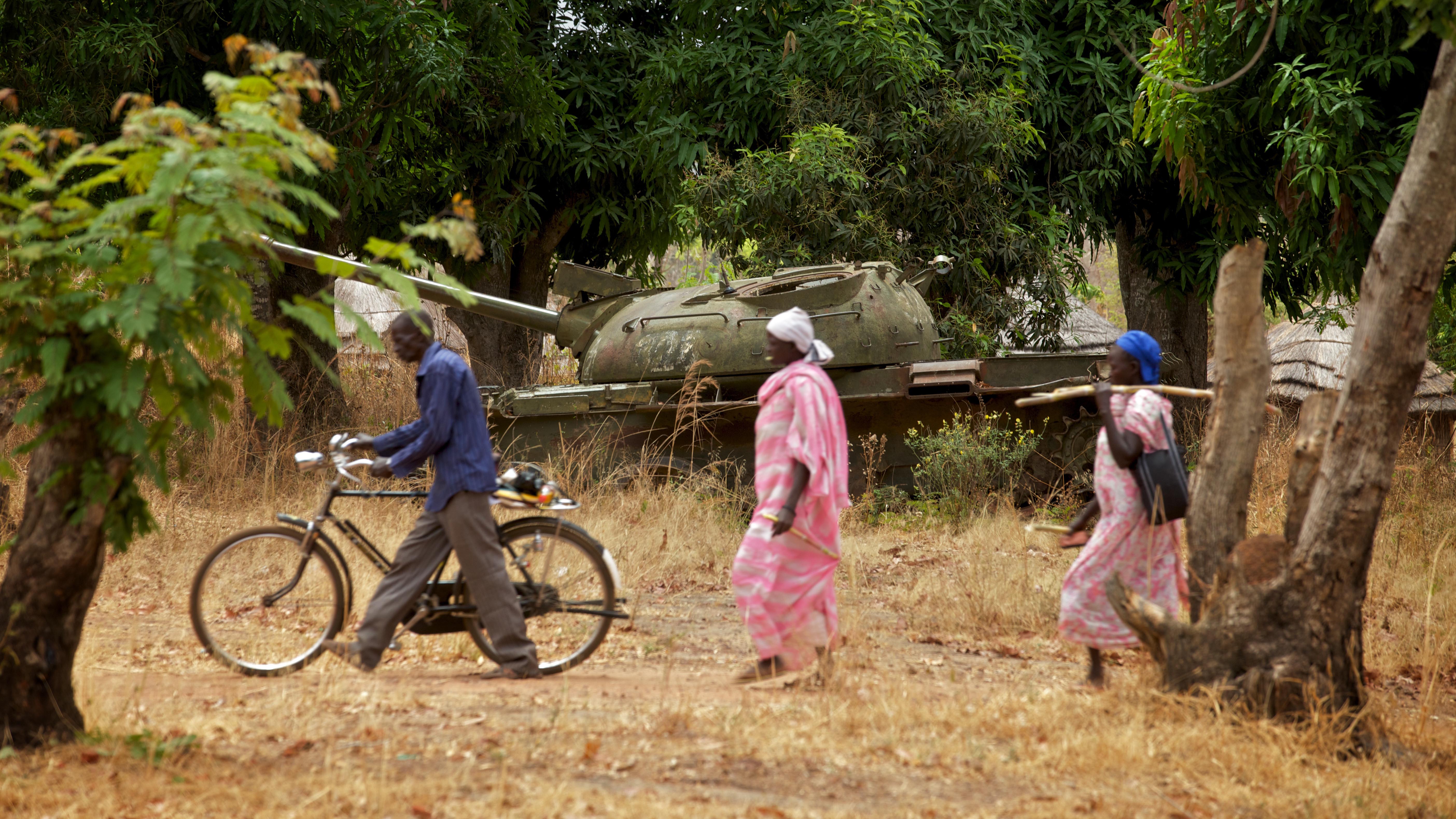 Africa - South Sudan - Community members walk past an old tank 