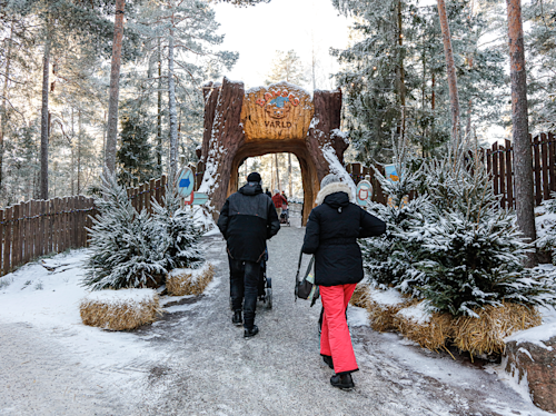 Människor går på en snöig stig genom en träbåge märkt "Värld" i ett skogsområde, omgiven av vintergröna träd och höbalar.
