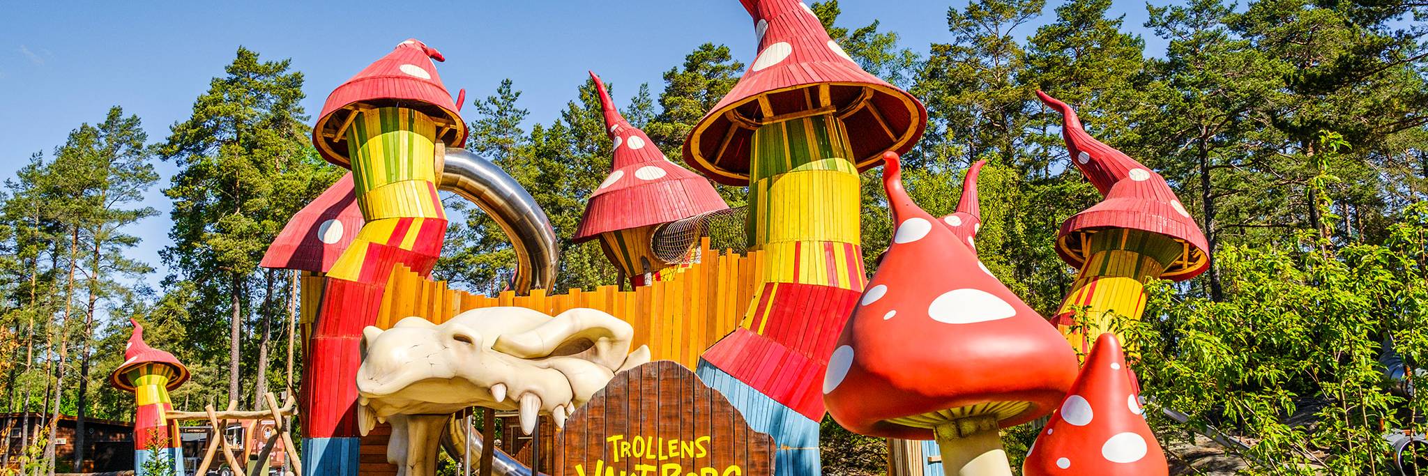 Färgglad och finurlig lekplats med röda svampformade torn, en stor djurskallemodell och en skylt med texten "Trollens Värld Borg", belägen i ett skogsområde under en blå himmel.