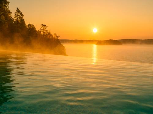 En infinitypool har utsikt över en lugn sjö i soluppgången, med dimma som stiger upp från vattnet och träd som avtecknar sig mot den orangefärgade himlen.