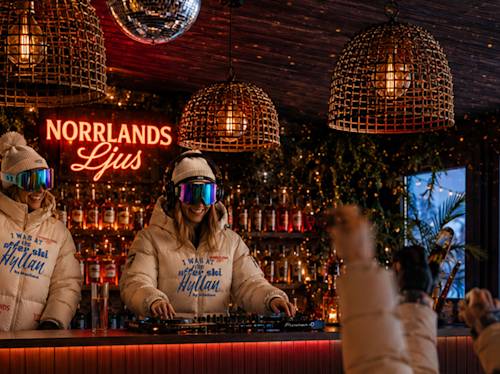 Two people in winter jackets and ski goggles stand at a bar with a DJ setup. Warm lighting, bottles on shelves, and a sign reading "NORRLANDS Ljus" are in the background.
