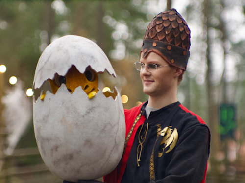 En person i kostym håller i ett stort, sprucket ägg som är en rekvisita, med en gul varelse inuti. Personen bär en medeltidsinspirerad dräkt och hatt. I bakgrunden syns träd.