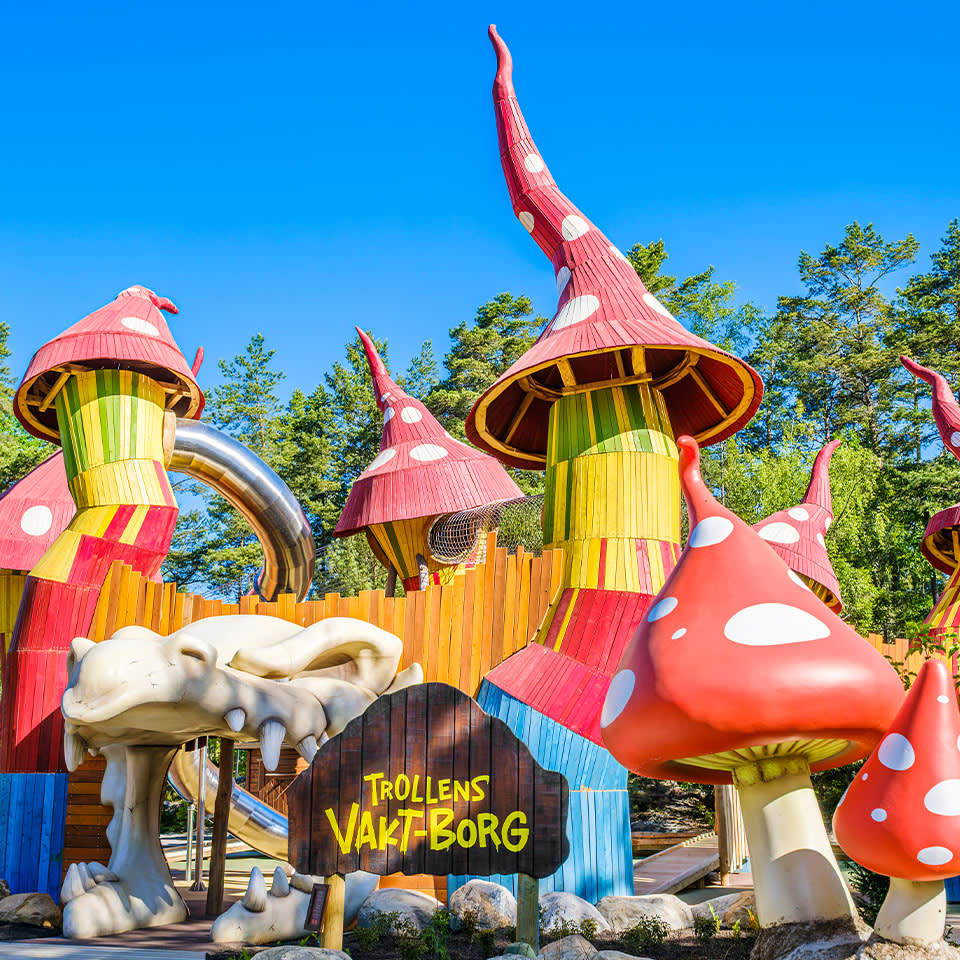 Färgglada, finurliga byggnader formade som höga, röda svampar med vita fläckar och en skylt med texten "Trollens Värltborg" på en utomhusnöjespark under en klarblå himmel.