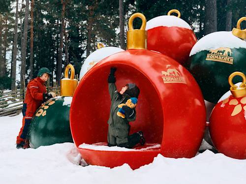 Två barn leker bland jättestora juldekorationer i en snötäckt utomhusmiljö; det ena barnet knäböjer inuti en röd juldekoration och håller i ett gosedjur, medan det andra står bredvid.