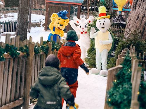 Två barn går på en snötäckt stig mot tre personer i djur- och karaktärskostymer som vinkar nära ett inhägnat område prytt med grönska.