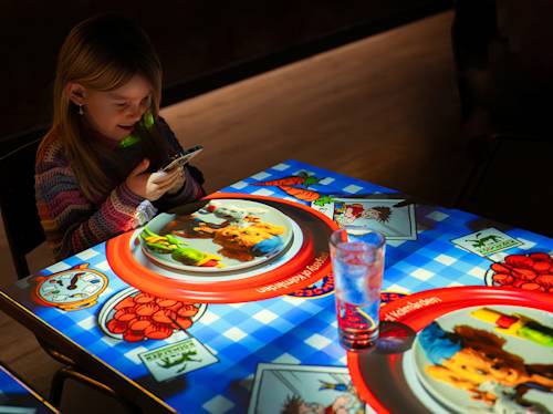 En ung flicka använder en smartphone vid ett upplyst interaktivt bord som visar animerade matbilder och lekfull grafik, med ett glas vatten i närheten.