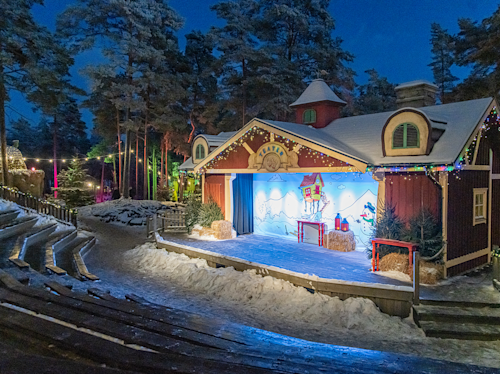 En vinterscen utomhus med festliga ljus och snöiga sittplatser, med en dekorerad bakgrund av ett hus och en snögubbe, omgiven av träd i skymningen.