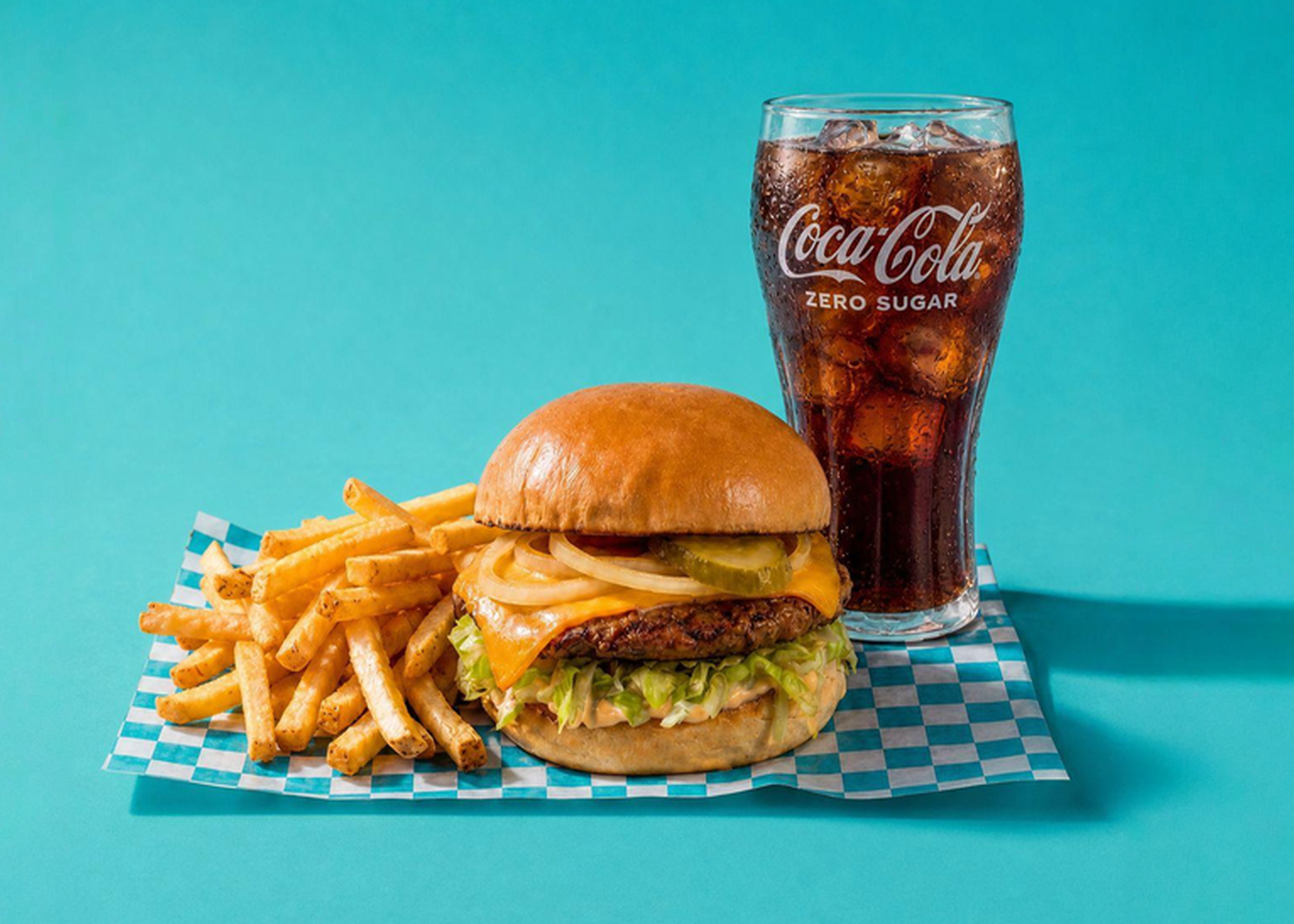 A cheeseburger with lettuce, tomato, and onions is served with French fries and a glass of Coca-Cola Zero Sugar on a blue checkered tray, against a plain blue background.