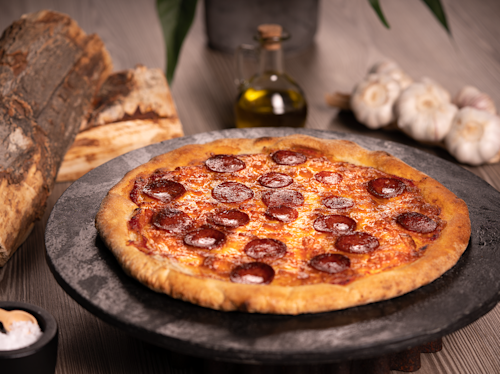 En tillagad pepperonipizza ligger på ett runt svart stenfat, med vitlök, olivolja och ved i bakgrunden på ett träbord.