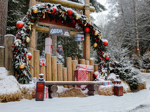 Stor julkrans med röda ornament dekorerar en träbåge märkt "Santa's Grotto". Inslagna julklappar och snötäckt mark kompletterar den festliga vinterscenen utomhus.