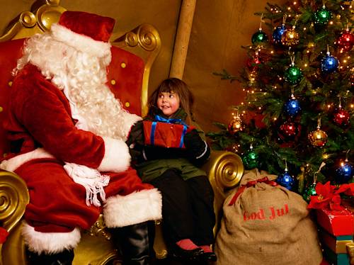 Ett barn sitter i jultomtens knä och får en inslagen present bredvid en pyntad julgran och en säck med texten ”God Jul”.