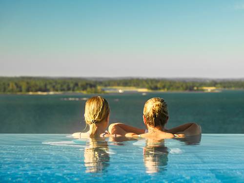 Två personer med blont hår kopplar av i en infinitypool med utsikt över en stor vattensamling och en grön strandlinje under en klar himmel.