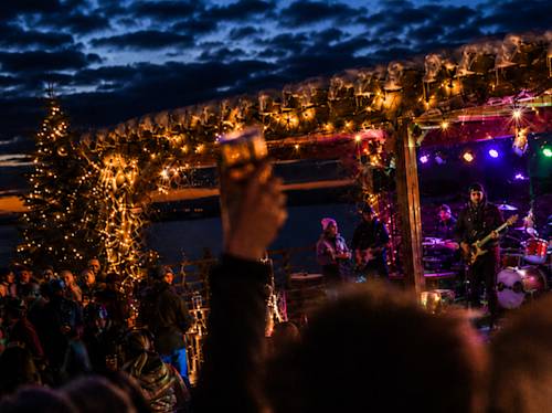 Outdoor evening concert with a lit stage, musicians performing, and holiday lights. A person in the crowd raises a drink; a decorated tree is visible in the background.