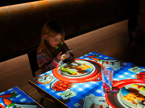 En ung flicka sitter vid ett bord med animerade matprojektioner medan hon tittar på en smartphone, med tomma glas och bestick på bordet.