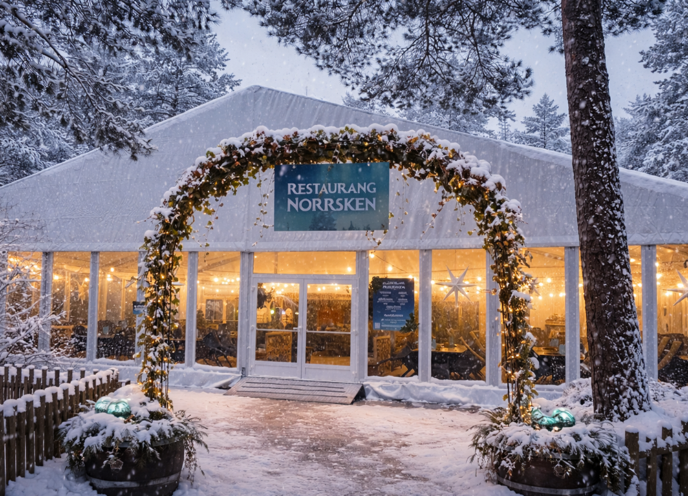 En snötäckt uteservering med namnet Restaurang Norrsken, med ljus som lyser inuti och en dekorerad båge vid entrén, som ligger bland tallar på vintern.