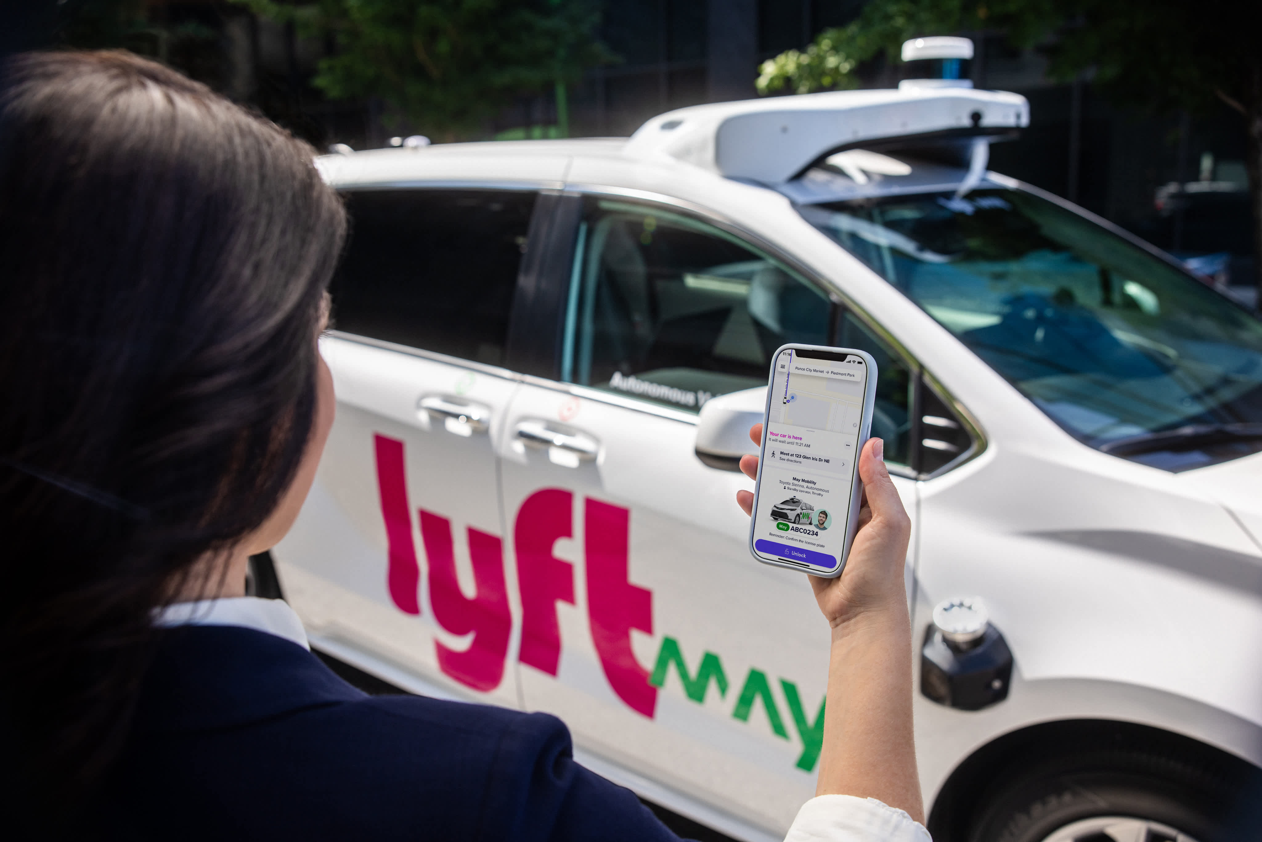 Person holding smartphone with Lyft app open next to white Lyft autonomous vehicle. 