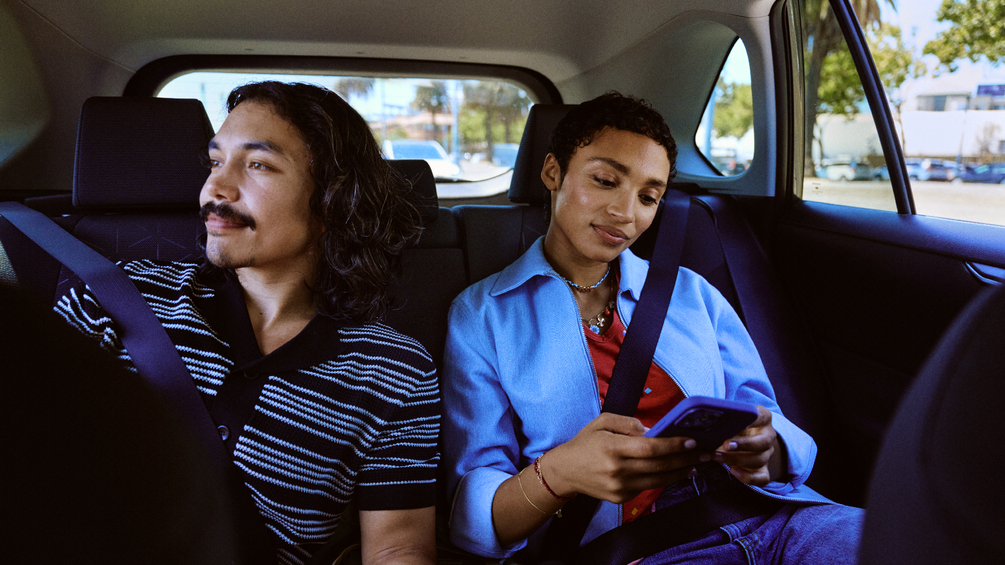 Two passengers in back seat of car, one looking out window while other checks phone. 
