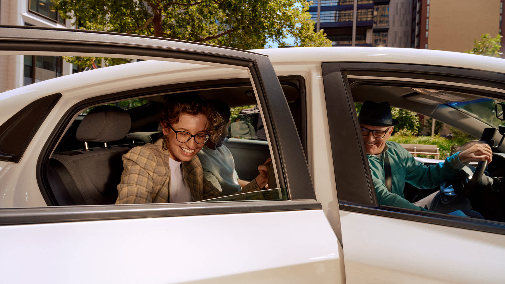 Passenger in plaid jacket smiling  with another passenger in a rideshare car with driver in blue shirt and hat.