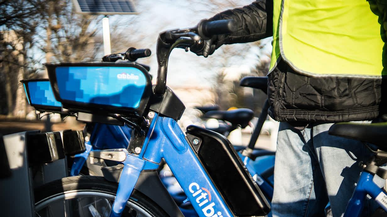 Photograph of a worker wearing a yellow safety vest putting a blue citibike bicycle into a dock rack.