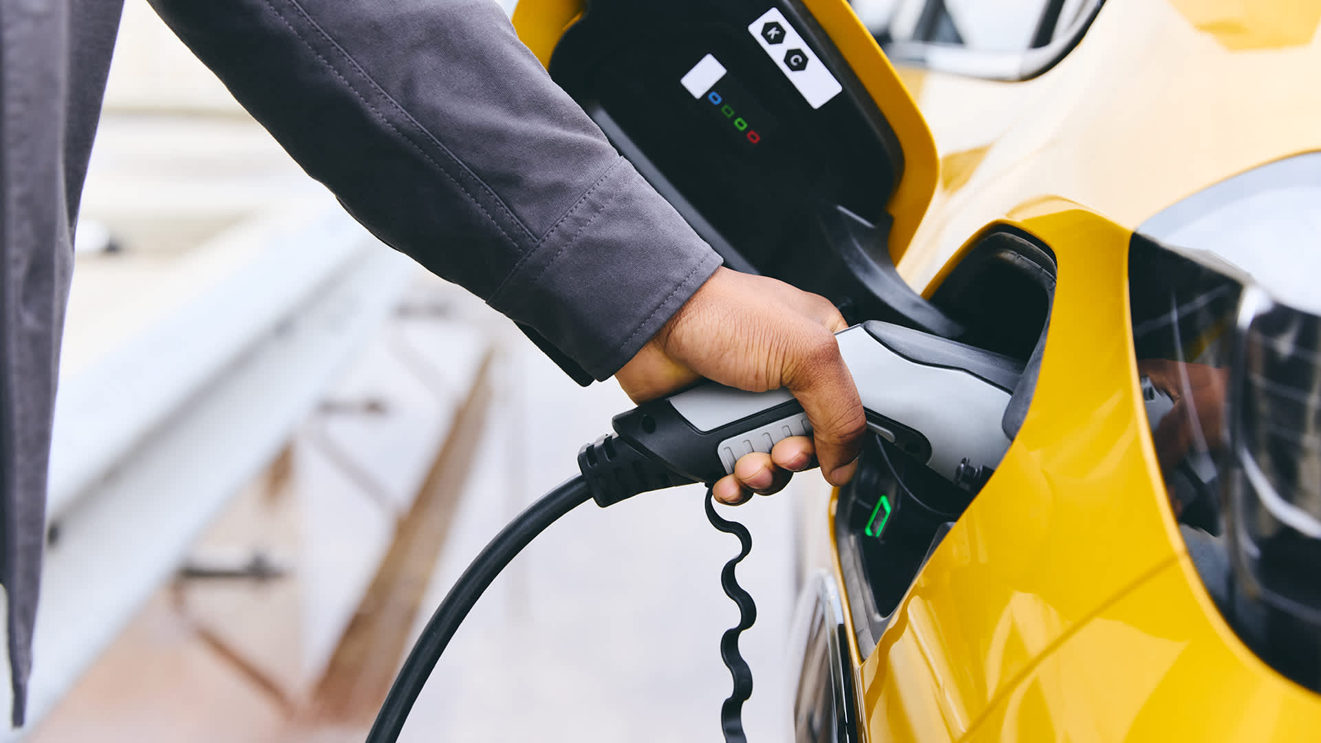 Photograph of a hand holding an electric vehicle charging plug in a yellow electric vehicle's charging port.